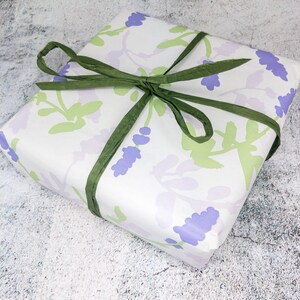 May include: A white gift box with a lavender floral pattern and green ribbon. The box is wrapped in a white paper with a lavender floral pattern and tied with a green ribbon.
