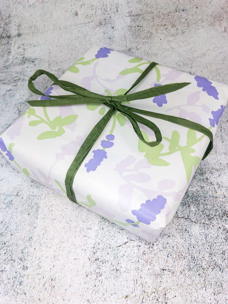 May include: A white gift box with a lavender floral pattern and green ribbon. The box is wrapped in a white paper with a lavender floral pattern and tied with a green ribbon.