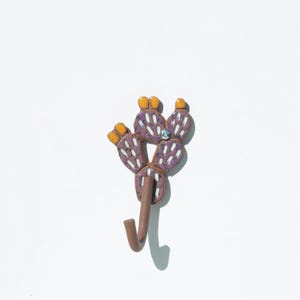 Könnte beinhalten: Lilafarbener und weißer Keramik-Kaktus-Haken mit einer gelben Blüte oben. Der Haken ist an einem braunen Metallstiel befestigt.