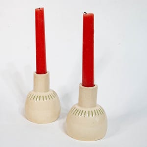 Puede incluir: Dos candeleros de cerámica con velas rojas. Los candeleros son de color blanco roto con una base redondeada y líneas verdes. Las velas rojas son altas y delgadas, creando un contraste llamativo con los tonos neutros.