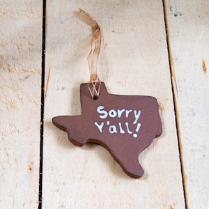 Può includere: Un ornamento in ceramica marrone a forma di Texas, con la scritta bianca "Sorry Y'all!". È appeso a un sottile nastro marrone chiaro. Lo sfondo è una superficie di legno chiaro. Il testo è in un carattere informale.