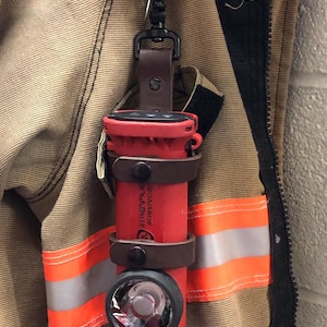 May include: A red flashlight with a black lens and a brown leather holster. The flashlight is attached to a black clip and has the text "SIREN" and "UL1950" printed on the side.