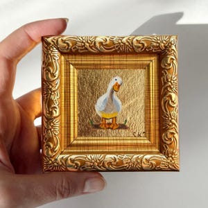 Op de afbeelding: Een kleine, vierkante schildering van een witte en grijze eend met oranje poten en snavel, tegen een gouden achtergrond. Het kunstwerk is omlijst in een sierlijke, goudkleurige lijst. De schildering wordt door een hand vastgehouden.