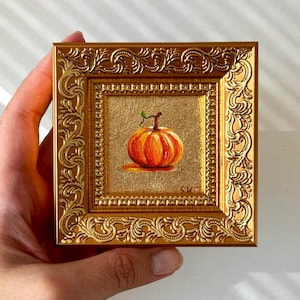 Puede incluir: Una pequeña pintura cuadrada de una calabaza naranja con un tallo verde centrada sobre un fondo dorado texturizado, encerrada en un marco dorado metálico ornamentado con intrincados patrones de volutas y las iniciales SKT en la esquina.