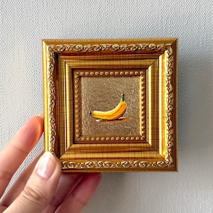 Peut inclure: Petite peinture d'une banane dans un cadre doré. La banane est jaune avec une tige verte, sur un fond doré. Le cadre est orné de sculptures détaillées et de forme carrée.