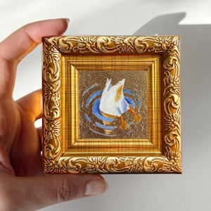 Op de afbeelding: Een kleine, vierkante schildering in een gouden, sierlijke lijst. Het kunstwerk toont een witte eend die in blauw water duikt. De lijst wordt door een hand vastgehouden. Afmetingen lijst: ca. 7,6 cm x 7,6 cm.