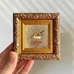 May include: A small, square, framed painting of a bird perched on a branch. The bird has yellow, green, and black feathers. The frame is ornate gold with a textured gold background. The painting is signed "SKJ".