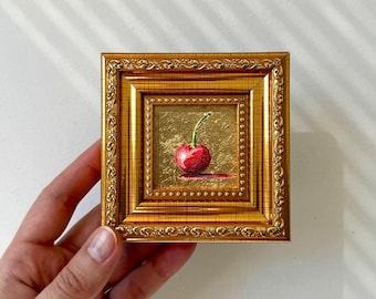 May include: A miniature painting of a single red cherry with a green stem, set against a gold background. The artwork is displayed in a small, ornate gold frame, held by a hand.