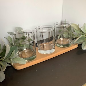 May include: Three clear drinking glasses with a black grid pattern are arranged on a light brown wooden tray. The glasses are cylindrical with a thick base. The tray is rectangular with rounded edges. Greenery is visible in the background.
