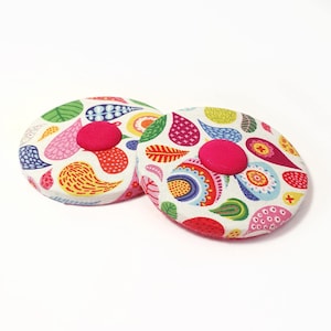 Könnte beinhalten: Zwei runde Stoffdeckel für Gläser mit einem Paisley-Muster in den Farben Rosa, Gelb, Grün, Blau und Rot. Jeder Deckel hat einen knallrosa Knopf in der Mitte.