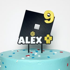 May include: A black and gold cake topper with the name "ALEX" and the number "9". The topper features a black square with a white square and gold accents, and is placed on a light blue cake with sprinkles.