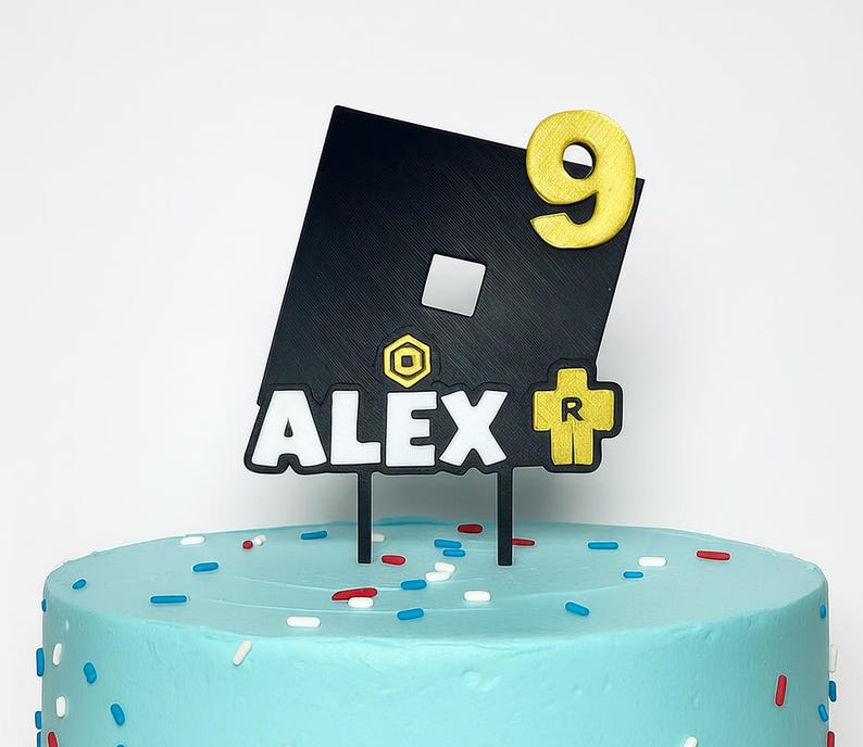 May include: A black and gold cake topper with the name "ALEX" and the number "9". The topper features a black square with a white square and gold accents, and is placed on a light blue cake with sprinkles.