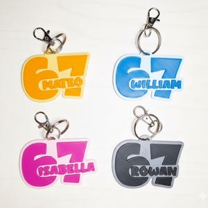 May include: Four personalised keychains in yellow, blue, pink, and grey. Each keychain features the number "67" with a name: Mateo, William, Isabella, and Rowan. Each keychain has a silver-coloured clasp.