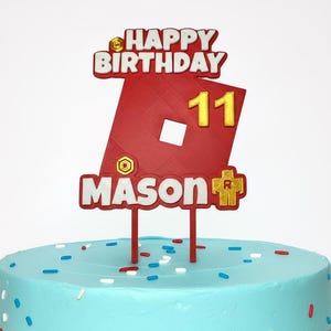 May include: A red and white cake topper with the words "HAPPY BIRTHDAY" and "MASON" in white lettering. The number "11" is in gold. The topper is on a blue cake with sprinkles.