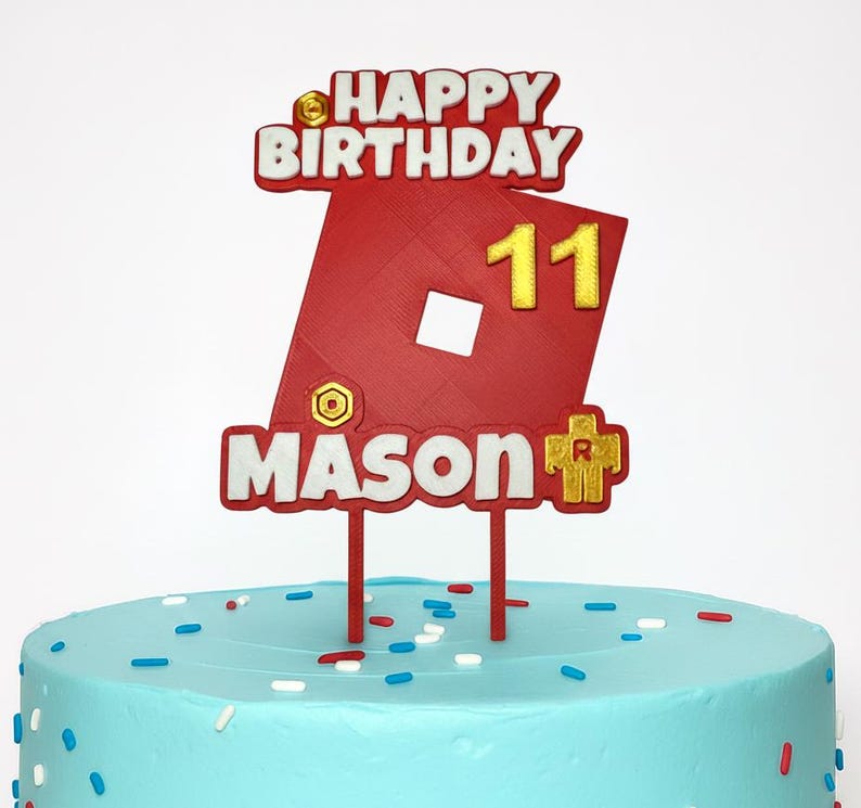 May include: A red and white cake topper with the words "HAPPY BIRTHDAY" and "MASON" in white lettering. The number "11" is in gold. The topper is on a blue cake with sprinkles.