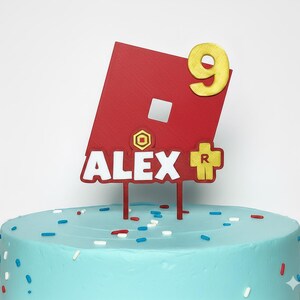 May include: A red and white cake topper with the name "ALEX" and the number "9" in gold. The topper features a red square with a white square and a yellow cross with the letter "R". The cake is light blue with sprinkles.