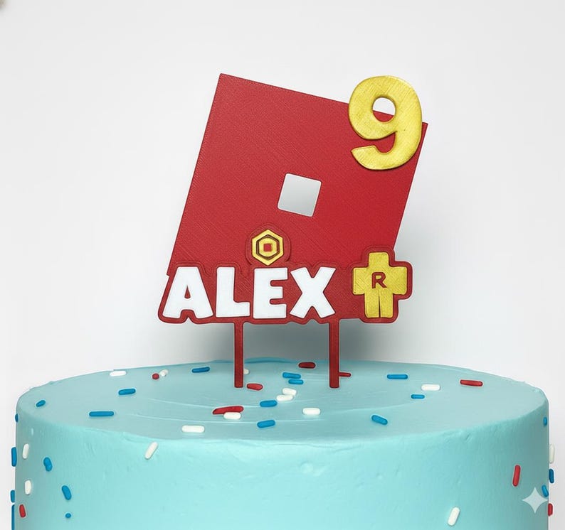 May include: A red and white cake topper with the name "ALEX" and the number "9" in gold. The topper features a red square with a white square and a yellow cross with the letter "R". The cake is light blue with sprinkles.