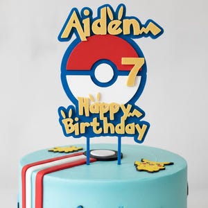 May include: A blue frosted cake with a decorative topper that says "Aiden 7 Happy Birthday". The topper features a red, white, and blue design, and the cake is decorated with yellow Pikachu figures.