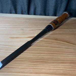 Op de afbeelding: Een Japanse houtbeitel met een lang, donker, rechthoekig metalen blad met een afgeschuinde rand. Het handvat is gemaakt van lichtbruin hout met een donkerblauwe ring en een metalen dop aan het uiteinde, voorzien van een klein, kleurrijk label.