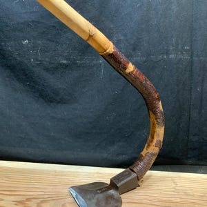 May include: A curved, antique hoe with a wooden handle and a metal blade. The handle is made of light-colored wood and is attached to a curved section wrapped in a darker material. The blade is a dark, rectangular shape.