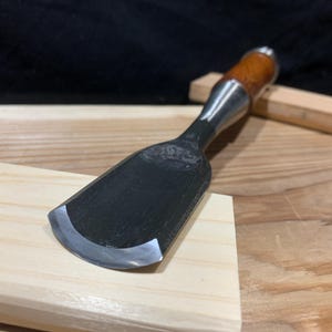 Peut inclure: Un gros plan d'un ciseau à bois avec un manche en bois brun et une lame tranchante et incurvée. Le ciseau repose sur un morceau de bois.