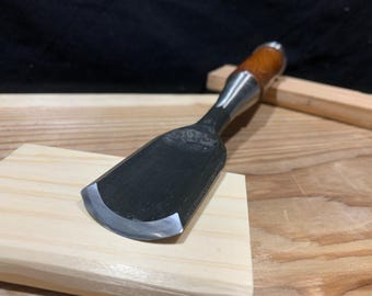 May include: A close-up of a wood carving chisel with a brown wooden handle and a sharp, curved blade. The chisel is resting on a piece of wood.