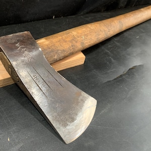 May include: A vintage axe with a wooden handle and a metal head. The axe head rests on a small wooden block. The handle is a light brown color, and the axe head is a darker, metallic color. The axe appears to be well-used.