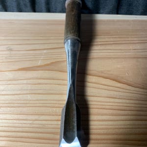 以下が含まれることがあります： 丸いキャップが付いたダークブラウンの木製ハンドルと、長いテーパー状のスチールシャフトを備えた金属製の木工用ノミ。シャフトは湾曲した凹型の刃へと続き、先端には鋭く磨かれた刃先が付いています。