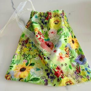 Puede incluir: Una bolsa de cordón verde con estampado floral que presenta girasoles, rosas y otras flores en tonos amarillos, rojos, rosas y azules. La bolsa tiene un cordón de cinta blanca.