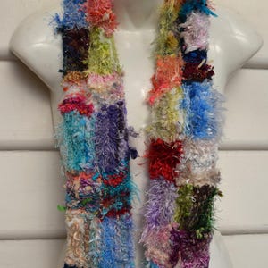 May include: A vibrant, handmade scarf on a mannequin. The scarf has a patchwork design with shades of blue, red, purple, and green. The scarf has a textured, fuzzy appearance, suggesting a soft, warm material, perfect for colder weather.
