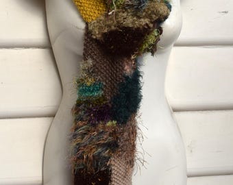 May include: A handmade, colorful scarf displayed on a mannequin. The scarf features a patchwork design with various textures and colors, including green, yellow, brown, and pink. The scarf is made of knitted and fuzzy materials.