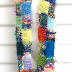 May include: A colourful patchwork scarf with a fuzzy texture. The scarf is made of different coloured yarns and is tied together in a knot.