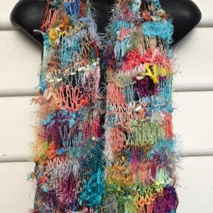 May include: A multi-coloured scarf featuring a complex, textured weave of various yarns, including fuzzy, metallic, and ribbon-like strands. The design incorporates a vibrant palette of blue, pink, yellow, orange, and purple hues in a loose, open-knit pattern.