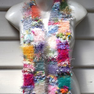 May include: A colorful, multi-textured scarf made of yarn in shades of pink, purple, blue, green, yellow, and white. The scarf is displayed on a white mannequin.