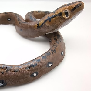 May include: A handcrafted, coiled snake sculpture. The snake features a brown body with black and white markings. The head is detailed with gold and black accents, and a realistic eye. The sculpture is set against a white background.