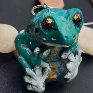 May include: A handcrafted frog pendant with a silver-colored bail. The frog is primarily teal with yellow and black eyes, and gray feet. The frog's belly has a blue and yellow iridescent pattern.