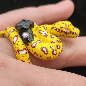 May include: A yellow snake ring with red and white spots and a black head. The snake is coiled around the finger, with the head resting on top. The ring is made of a painted material.
