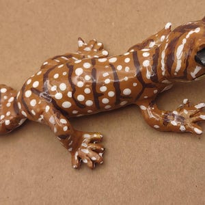 May include: A brown and white spotted gecko figurine with dark brown stripes. The lizard has detailed claws and a realistic eye. The gecko is a decorative item, possibly for a shelf or desk.