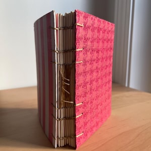 May include: A pink and white striped book with a pink patterned cover. The book is bound with a brown thread and has a spine made of natural material.