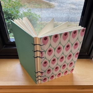 May include: A handmade journal with a green striped cover and a pink and white ikat patterned cover. The journal is bound with black thread and has a spine made of wood.