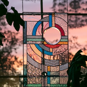 May include: A stained glass window hanging in a window frame. The window is made of colorful glass pieces in geometric shapes, including a circle in the center. The background is a blurry sunset.