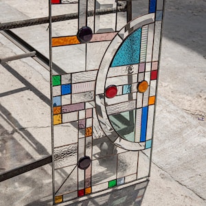 May include: A rectangular stained glass panel featuring a geometric composition of clear textured glass panes interspersed with small squares and circles in red, blue, orange, purple, and green, framed by thin metal cames in a grid pattern.