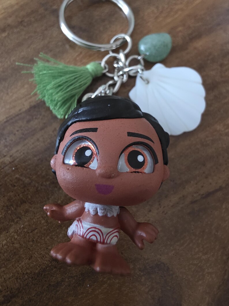 May include: A keychain featuring a small figurine of a cartoon character with brown skin, black hair, and large eyes. The keychain includes a green tassel, a white seashell charm, and a green stone. The figurine is wearing a white and red patterned garment.