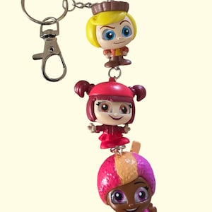 May include: A keychain featuring three colorful cartoon character figurines. The top figurine has blonde hair and a brown hat. The middle figurine is wearing a red outfit. The bottom figurine has pink and purple hair and a pink outfit.