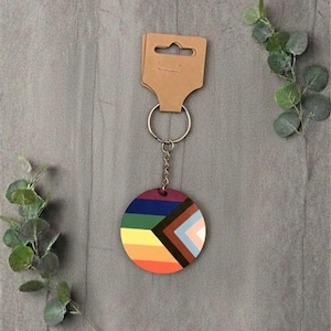 May include: A circular keyring featuring a rainbow design with a brown and black geometric pattern. The keyring is attached to a silver metal ring and chain, displayed on a grey surface.