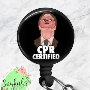 Puede incluir: Un portatarjetas redondo de color negro con una ilustración de dibujos animados de una persona con una máscara facial de material color piel, vistiendo un traje marrón y corbata. Debajo, el texto blanco dice CPR CERTIFIED en mayúsculas.