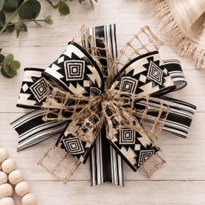 May include: A decorative bow crafted from multiple ribbons, featuring a black and white geometric diamond pattern, black and white stripes, and a natural tan jute mesh netting. The ribbons are layered and tied together in a central knot.