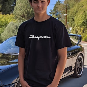 May include: A black Toyota Supra sports car with a young person standing next to it. The person is wearing a black t-shirt with the word "Supra" printed in white on the front. The car is parked on a paved driveway.
