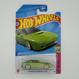 May include: A green '89 Mazda Savanna RX-7 FC3S die-cast toy car on a blue and red Hot Wheels card. The car has gold-colored wheels and is part of the HW: The '90s series. The card features the Hot Wheels logo and a guarantee for life.