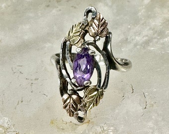 Puede incluir: Un anillo de plata con una amatista púrpura engastada en un diseño de hojas. El anillo está adornado con hojas doradas.